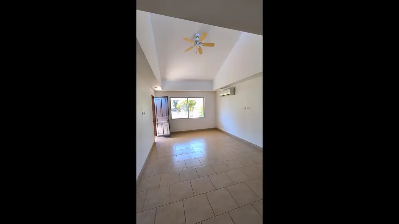 Long interior view from the kitchen side toward the front window, open door, ceiling fan, and wall AC unit